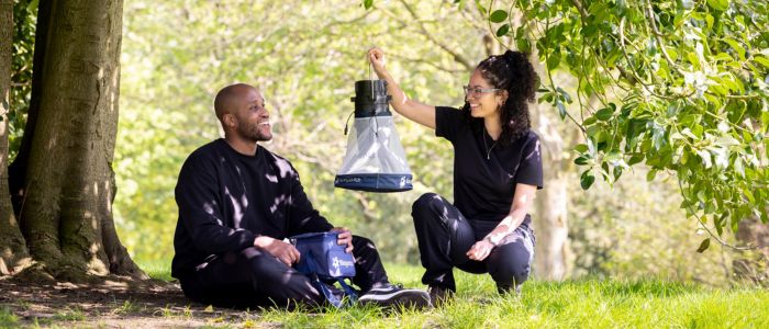 One male and one female sat on the floor outdoors surrounded by trees and grass, holding a small mosquito net.