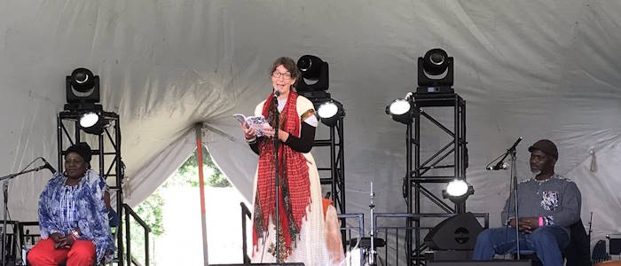 Alison Phipps standing on stage reading from a book with Gameli and Naa Densua Tordzro seated on either side of her