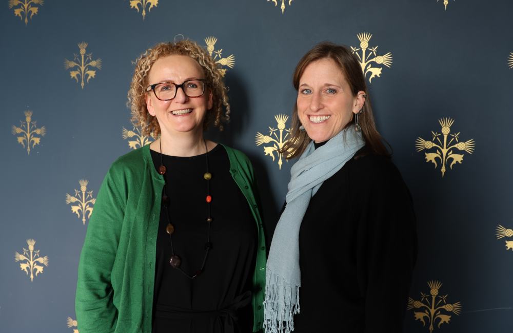 Two people standing next to each other smiling against a wall with blue wallpaper