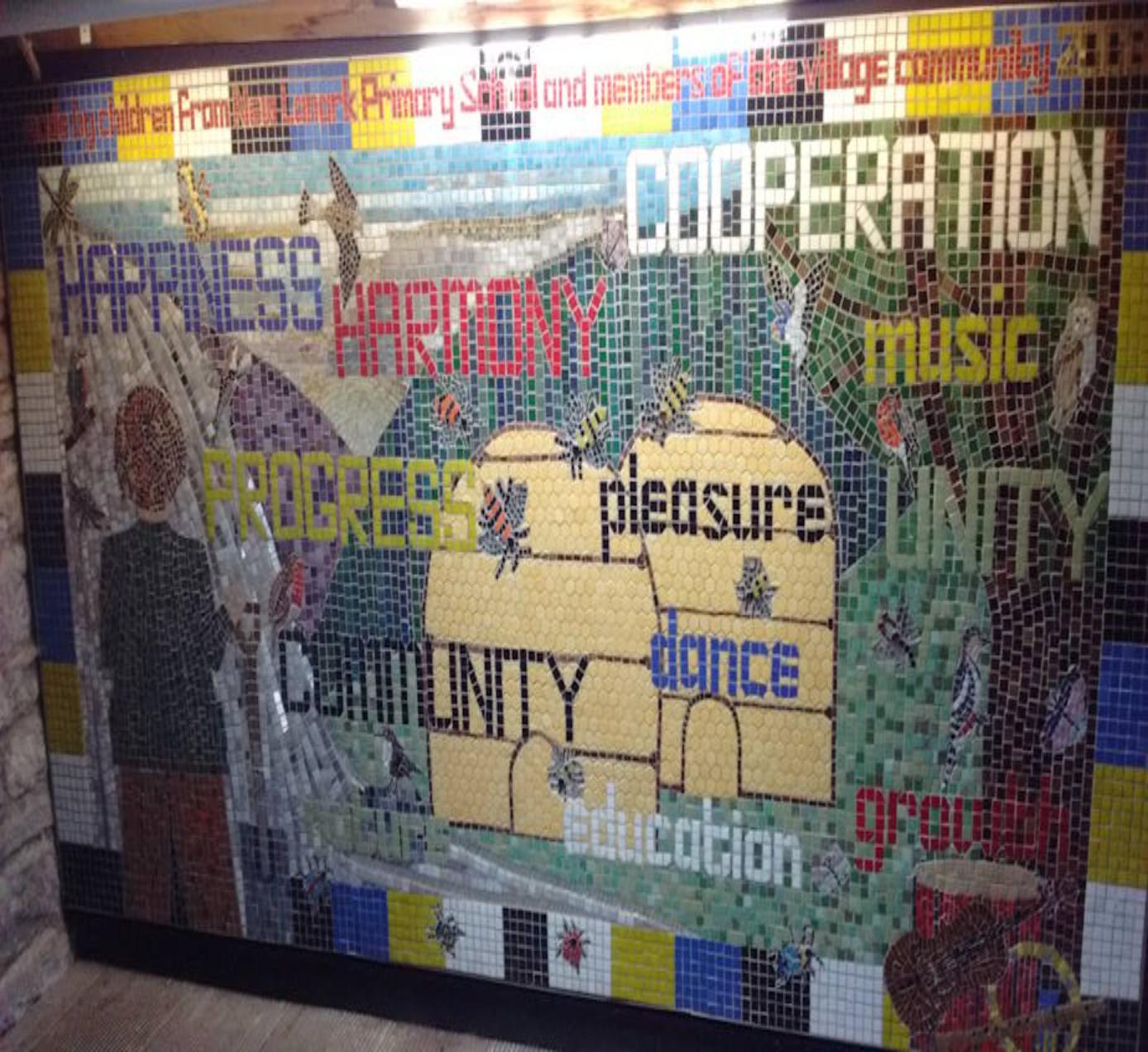 Colourful mosaic made out of small tiles depicting a person with their back to the viewer looking out toward a hilly view with beehives, a tree, bees, other insects and birds in the foreground. There are words in different colours scattered across the mosaic: happiness, harmony, cooperation, music, progress, pleasure, unity, community, dance, education, growth.