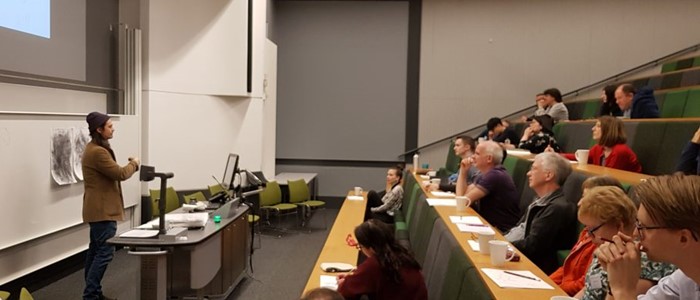 a panoramic view of an auditorium with a person in in foreground, in front of a projection screen and of mixed-gender members of the audience 