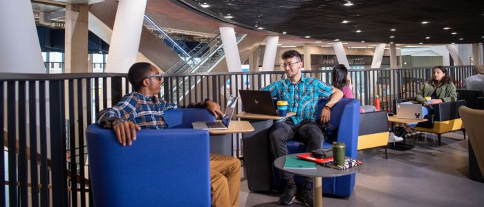 Two students sat talking with laptops on tables inside the James McCune Smith Learning Hub