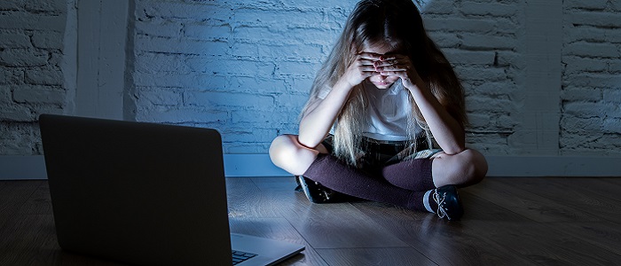 A young girl sitting crosslegged on the floor with her head in her hands, lit by the glow from the open laptop on the the floor beside her. Source: SB Arts Media | iStockphoto https://www.istockphoto.com/photo/scared-sad-girl-bullied-on-line-with-laptop-suffering-cyberbullying-and-harassment-gm964741476-263339312