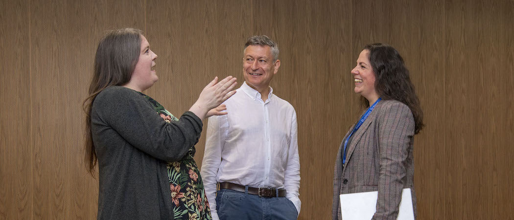 A woman gesturing as she speaks to a man and woman who are smiling as they listen. They are all standing up
