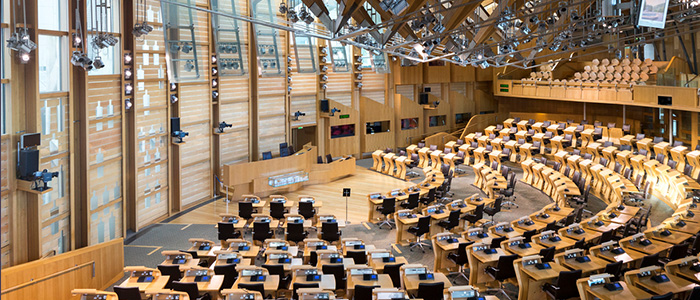 The Scottish Parliament chamers with multiple seats and desks for every MSP in the region. In addition sits the prosiding chair facing the other desks present. The room is empty and primarily has wooden based features on the walls and the floor