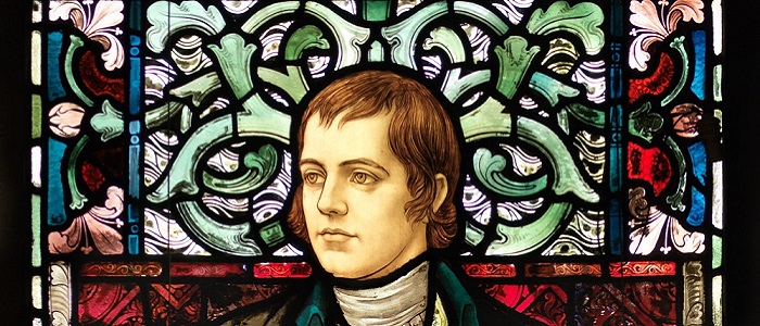 Stained glass window with a portrait of Robert Burns on a floral background and the name 'Burns' in the window above his head. Source: University of Glasgow https://www.gla.ac.uk/events/globalburns/