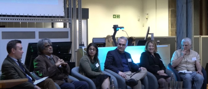 Panel of six speakers seated in front of an audience, and conversing with each other