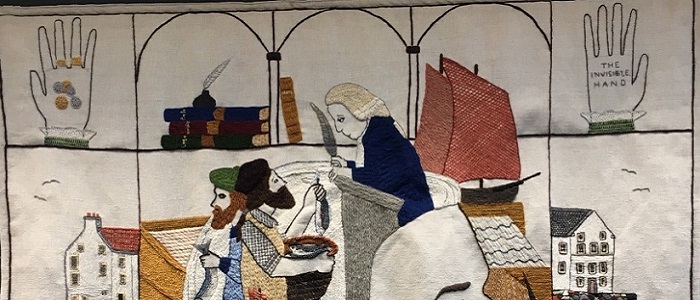 Part of an embroidered panel in Great Tapestry of Scotland depicting Adam Smith at a desk with a quill as fishermen hold up fish to him. Above him is pile of books, and a hand holding coins, and an empty hand called the Invisible Hand. To the side is a sailing boat and old houses. Source: L Janes