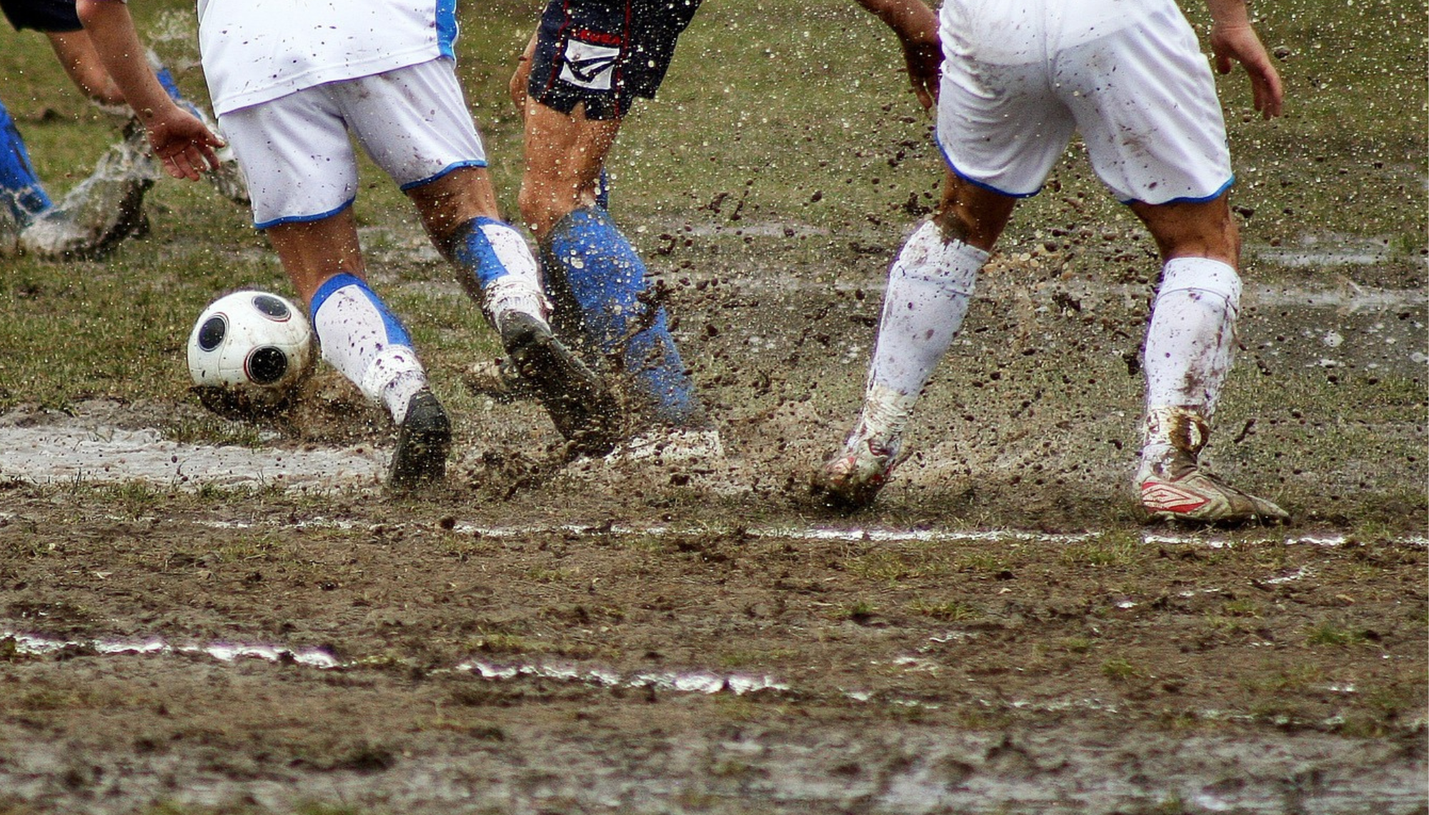 The image depicts mens legs playing football, lots of mud is splashing everywhere. They are wearing football shorts and shoes.